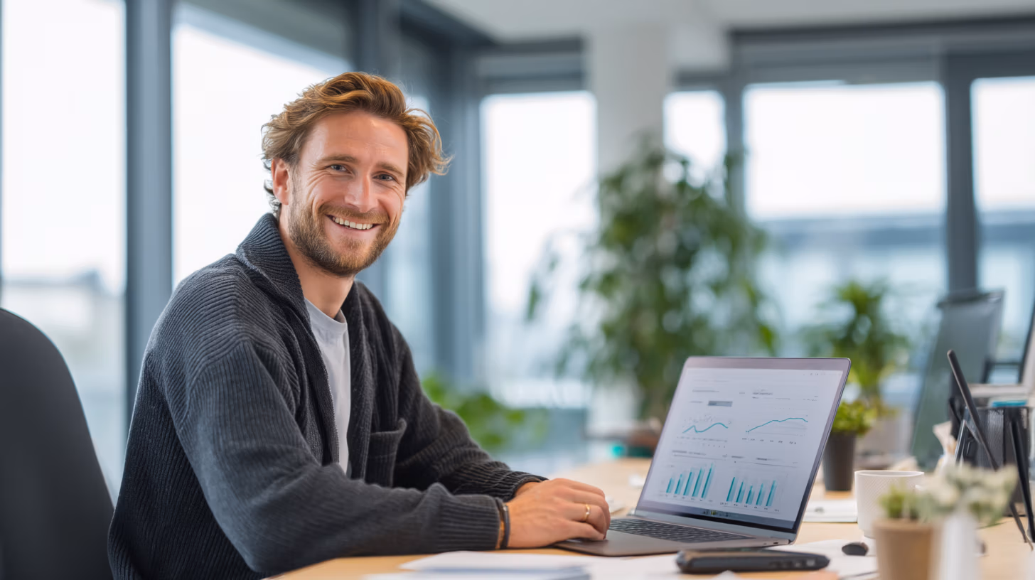 Lächelnder junger Mann im modernen Büro vor Laptop mit Verkaufsdiagrammen